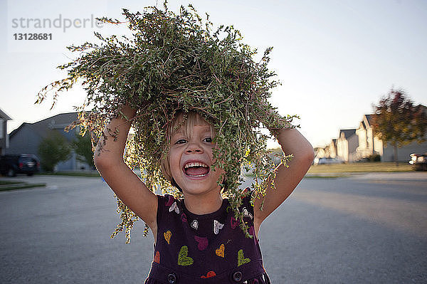 Porträt eines glücklichen Mädchens  das Pflanzen auf dem Kopf trägt  während es auf der Straße gegen den Himmel läuft
