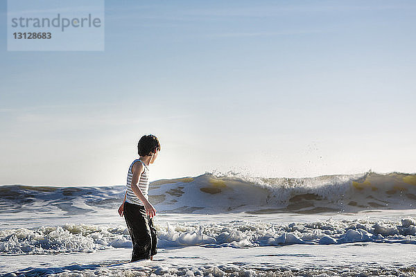 Seitenansicht eines Jungen  der mit Wellen spielt  während er bei Sonnenuntergang im Meer gegen den Himmel steht
