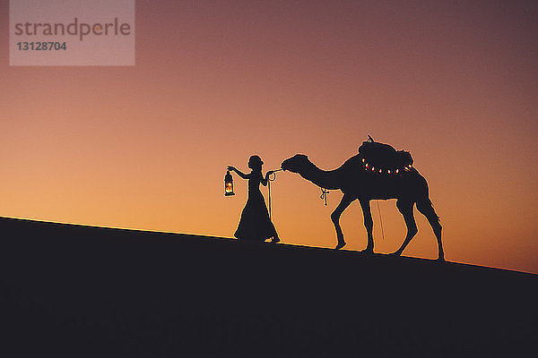 Scherenschnittfrau mit Kamel beim Spaziergang in der Sahara vor klarem Himmel bei Sonnenuntergang