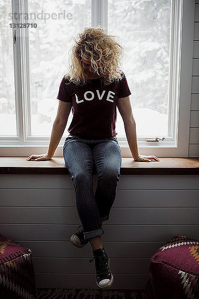 Frau in voller Länge trägt ein T-Shirt mit dem Text LOVE  während sie zu Hause auf dem Fensterbrett sitzt