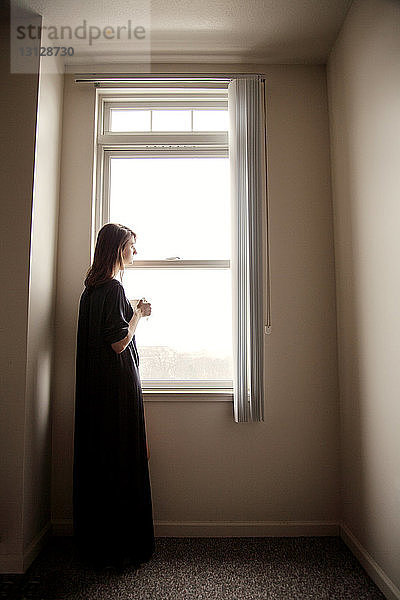 Seitenansicht einer Frau  die weg schaut  während sie zu Hause am Fenster steht