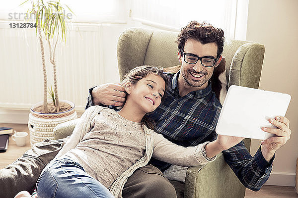 Liebevoller Vater und Tochter schauen auf das digitale Tablet  während sie auf einem Stuhl sitzen