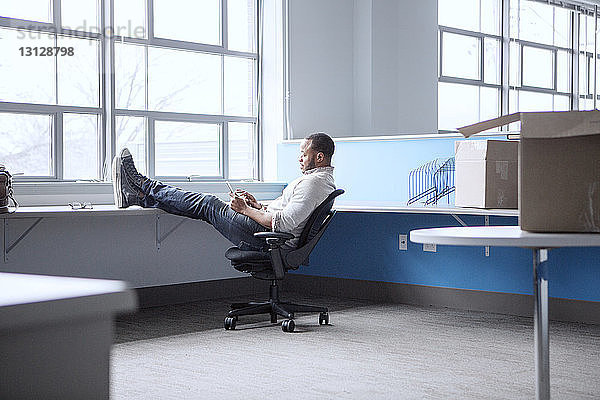 Geschäftsmann benutzt Mobiltelefon  während er sich im Büro auf einem Stuhl entspannt