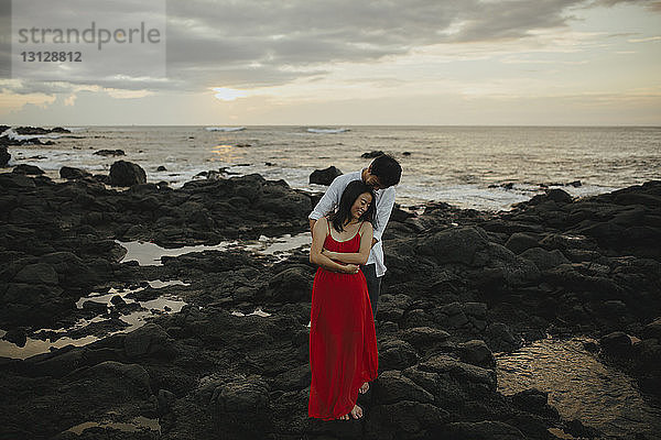 Glückliches Paar umarmt sich  während es bei Sonnenuntergang am Strand gegen den Himmel steht