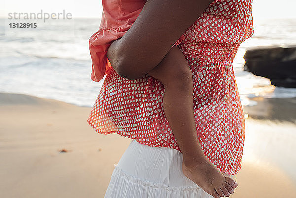 Mitschnitt einer Mutter  die eine Tochter trägt  während sie bei Sonnenuntergang am Strand steht