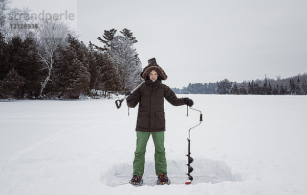 Porträt eines Mannes  der auf einem zugefrorenen See stehend Eisbohrer und Schaufel gegen den Himmel hält