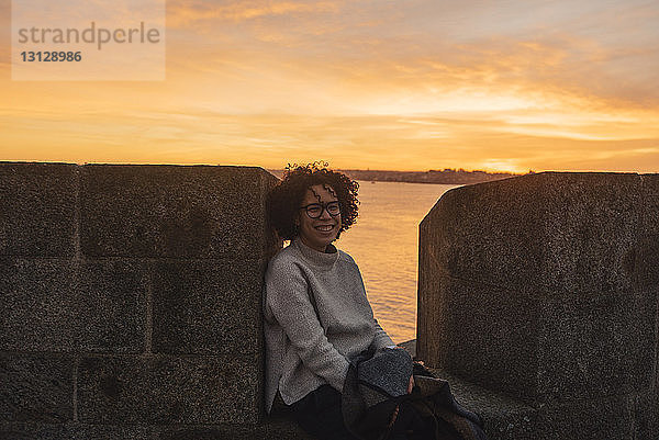 Fröhliche Frau sitzt bei Sonnenuntergang auf der Stützmauer eines historischen Gebäudes gegen das Meer