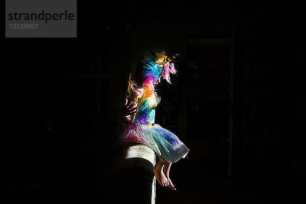 Seitenansicht eines Mädchens in farbenfrohem Einhorn-Kostüm  das zu Hause in der Dunkelkammer auf dem Sofa sitzt
