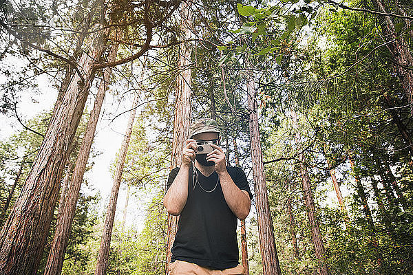 Mann fotografiert durch eine Kamera  während er im Wald an Bäumen steht