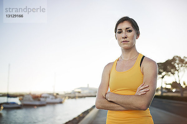Porträt einer selbstbewussten Sportlerin  die mit verschränkten Armen am Hafen vor klarem Himmel steht