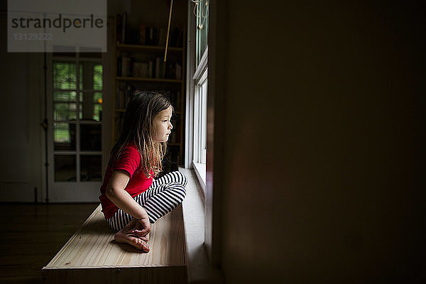 Seitenansicht eines niedlichen Mädchens  das durch ein Fenster schaut  während es zu Hause auf einem Holztisch sitzt