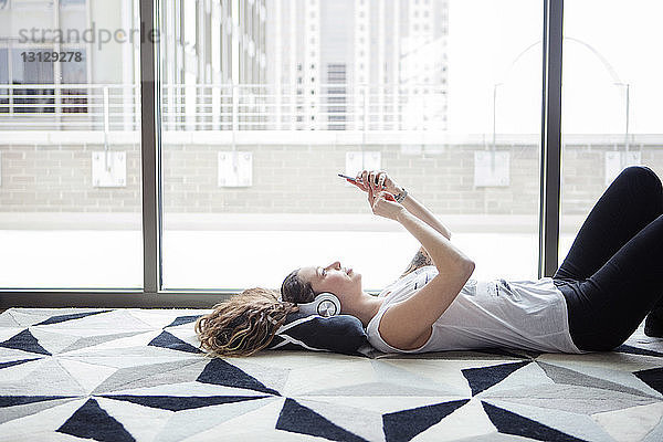 Seitenansicht einer jungen Frau  die ein Smartphone benutzt  während sie zu Hause am Fenster auf dem Teppich liegt