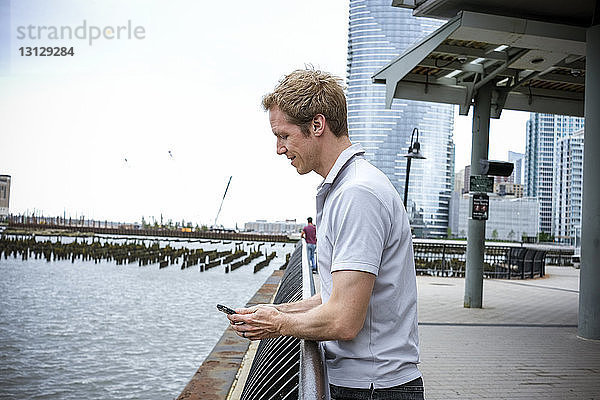Seitenansicht eines Mannes  der ein Smartphone benutzt  während er am Fluss auf der Promenade gegen den Himmel in der Stadt steht
