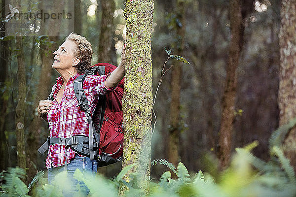 Glückliche ältere Frau mit Rucksack an Bäumen im Wald stehend
