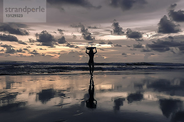 Silhouette einer jungen Frau  die bei Sonnenuntergang am Strand vor bewölktem Himmel ein Surfbrett auf dem Kopf trägt