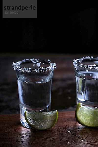 Nahaufnahme von Tequila mit Limettenscheiben auf Holztisch vor schwarzem Hintergrund