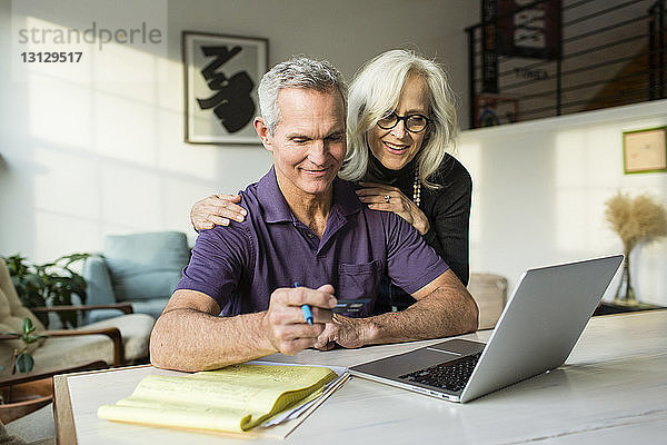 Lächelnde Frau schaut auf Laptop-Computer  während der Mann mit Kreditkarte im Wohnzimmer sitzt