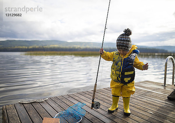 Ein Junge in voller Länge trägt Regenmantel und Schwimmweste  während er die Angelrute auf dem Holzsteg über dem See hält