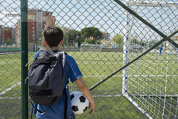 Rückansicht eines Jungen mit Rucksack  der einen Fussball hält  während er am Zaun am Unterstand steht