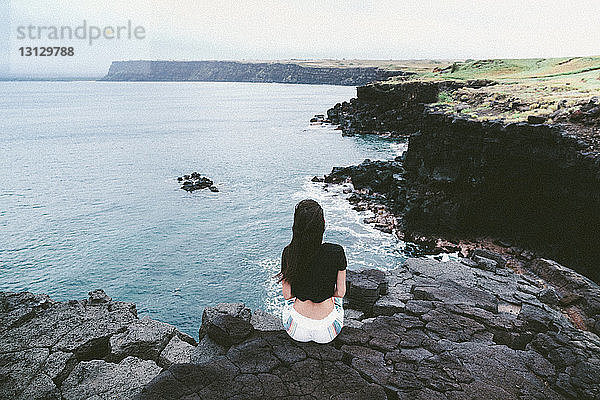 Rückansicht einer auf Felsen am Strand sitzenden Frau gegen den Himmel