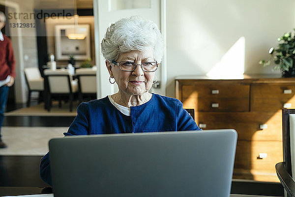 Ältere Frau benutzt zu Hause einen Laptop-Computer mit einem älteren Mann im Hintergrund