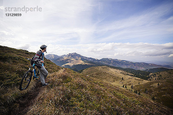 Männlicher Rucksacktourist lehnt mit dem Fahrrad auf grasbewachsenem Berg gegen den Himmel