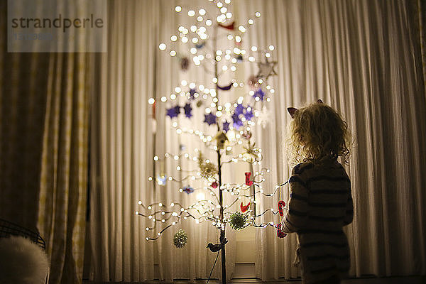 Rückansicht eines Mädchens  das zu Hause an einem künstlich beleuchteten Weihnachtsbaum vor Vorhängen steht