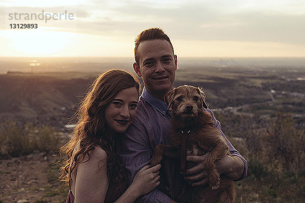 Porträt eines jungen Paares mit Hund  das bei Sonnenuntergang auf dem Feld steht