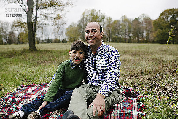 Porträt eines Vaters mit Sohn auf einer Decke gegen den Himmel im Park sitzend
