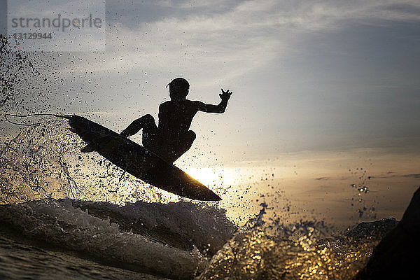 Junger männlicher Surfer springt bei Sonnenuntergang über das Meer