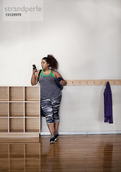 Frau in voller Länge  die ein Smartphone benutzt  während sie im Fitnessstudio an der Wand steht