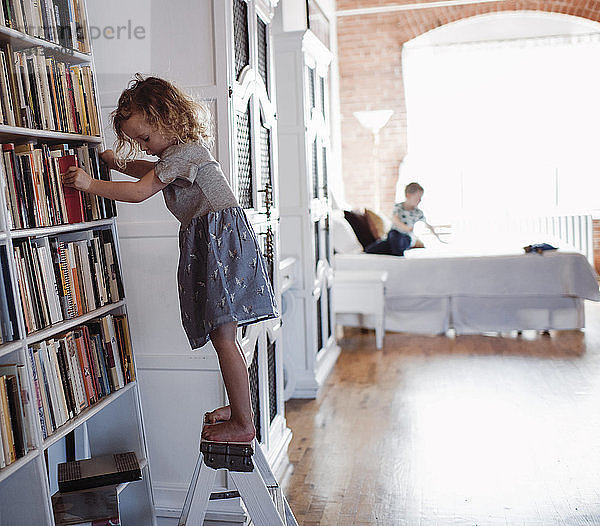 Seitenansicht eines Mädchens  das an Bücherregalen auf einer Leiter steht  mit dem Bruder im Hintergrund zu Hause