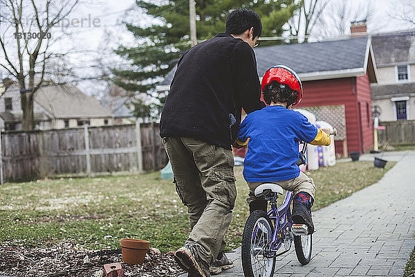 Rückansicht eines Vaters  der seinem Sohn beim Fahrradfahren auf einem Fußweg im Hinterhof hilft