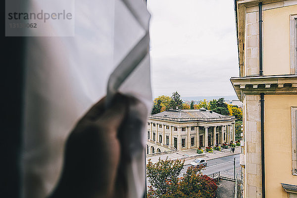 Abgehackte Hand eines Mannes  der einen Vorhang am Fenster gegen Gebäude hält