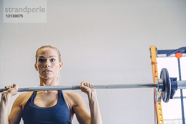 Selbstbewusste junge Frau hebt Langhantel gegen die Wand im Fitnessstudio