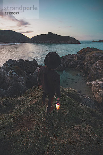 Rückansicht eines Mannes  der eine beleuchtete Laterne hält  während er bei Sonnenuntergang auf einem Felsen am Strand gegen den Himmel steht