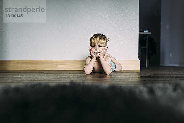 Porträt eines Jungen ohne Hemd mit Händen am Kinn  der zu Hause auf dem Boden liegt