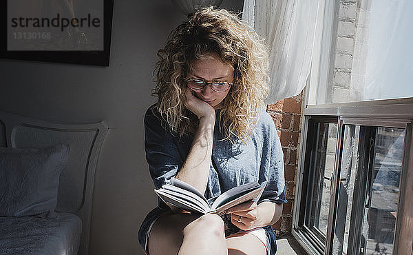Frau liest Buch  während sie zu Hause auf dem Fensterbrett sitzt