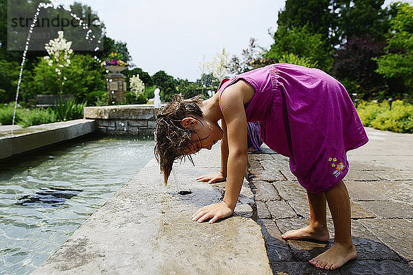 Seitenansicht eines verspielten Mädchens  das sich an einem sonnigen Tag im Park über einen Springbrunnen beugt