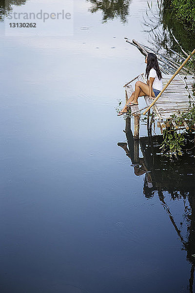 Hochwinkelansicht einer nachdenklichen Frau  die auf einem Pier am Fluss sitzt