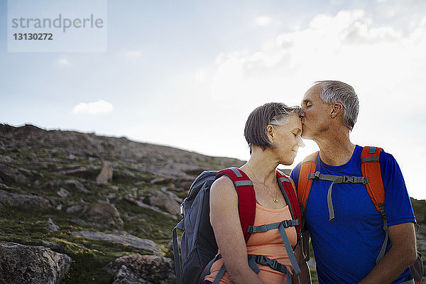 Ehemann küsst Frau auf die Stirn  während er auf Berg gegen Himmel steht