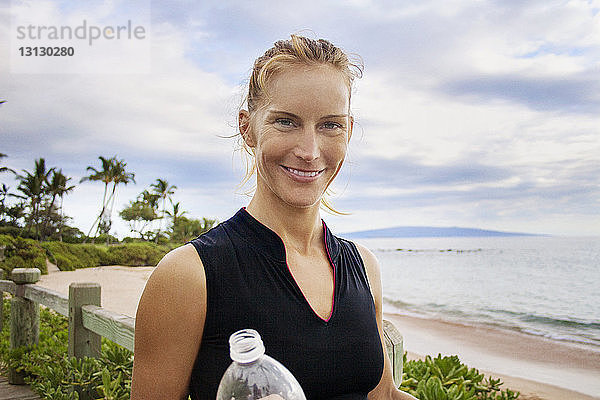 Porträt einer lächelnden Frau mit Flasche am Strand vor bewölktem Himmel