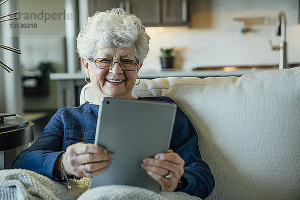Ältere Frau lächelt  während sie einen Tablet-Computer auf dem Sofa zu Hause benutzt