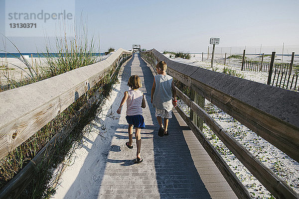 Rückansicht von Geschwistern  die bei klarem Himmel am Strand auf der Strandpromenade spazieren gehen