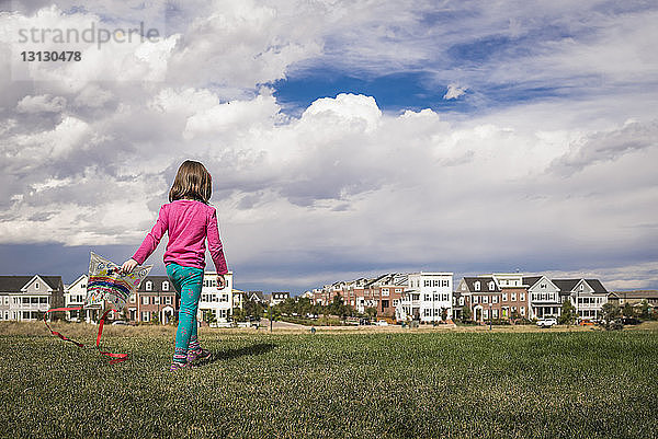 Rückansicht eines Mädchens mit Drachen  das auf einem Grasfeld vor Stadtbild und bewölktem Himmel läuft