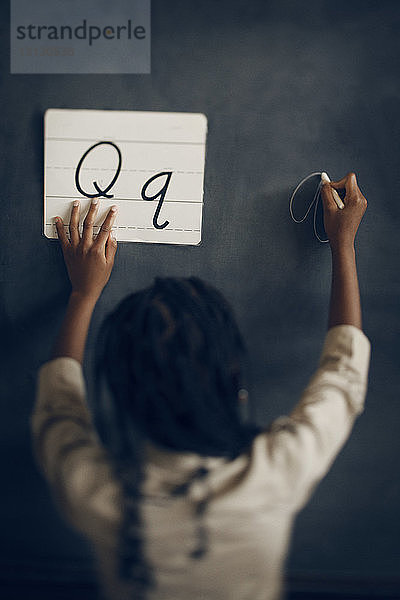 Rückansicht eines Schulmädchens  das das Alphabet Q schreibt  während es Papier an der Tafel hält