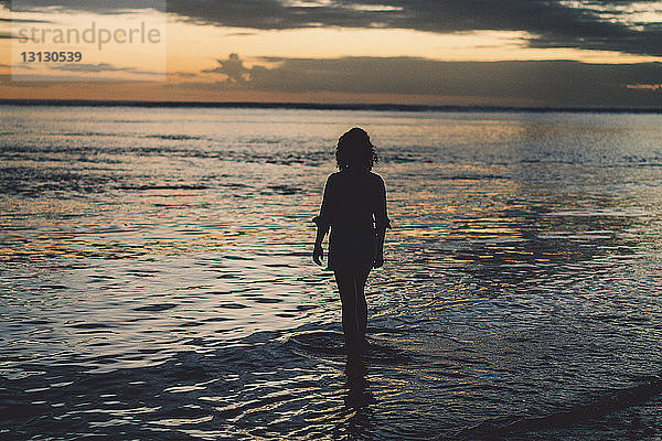 Silhouettenfrau steht am Strand vor bewölktem Himmel bei Sonnenuntergang