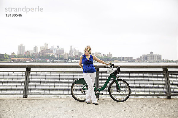 Porträt einer glücklichen  reifen Frau  die mit einem Fahrrad auf einer Promenade in der Stadt vor klarem Himmel steht