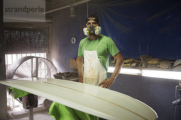 Porträt eines Arbeiters in Arbeitsschutzkleidung  der in der Werkstatt am Surfbrett steht