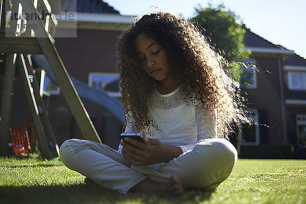 Mädchen benutzt Mobiltelefon  während sie auf einem Grasfeld im Hinterhof sitzt